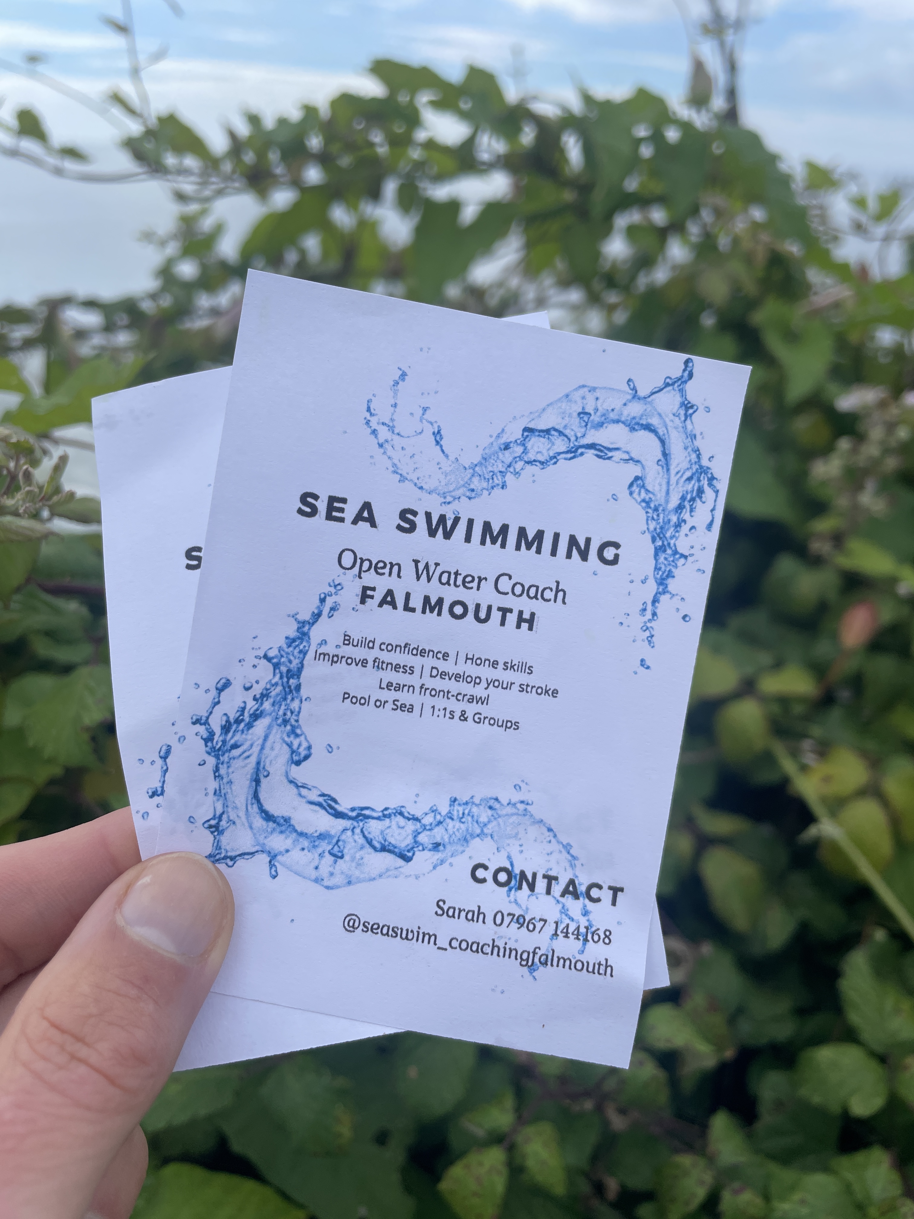 Image shows photo of hand holding two paper fliers promoting Sea Swim Coaching Falmouth, with contact details, sea and leafy foliage in the background.