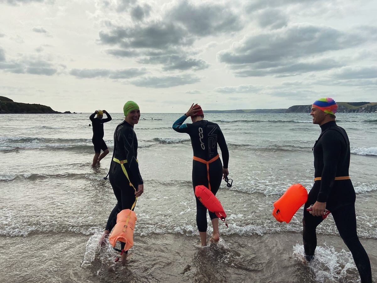 sea swimmers wearing swim wetsuits and tow floats entering the shallows before a swim, location south devon, clouds in the sky