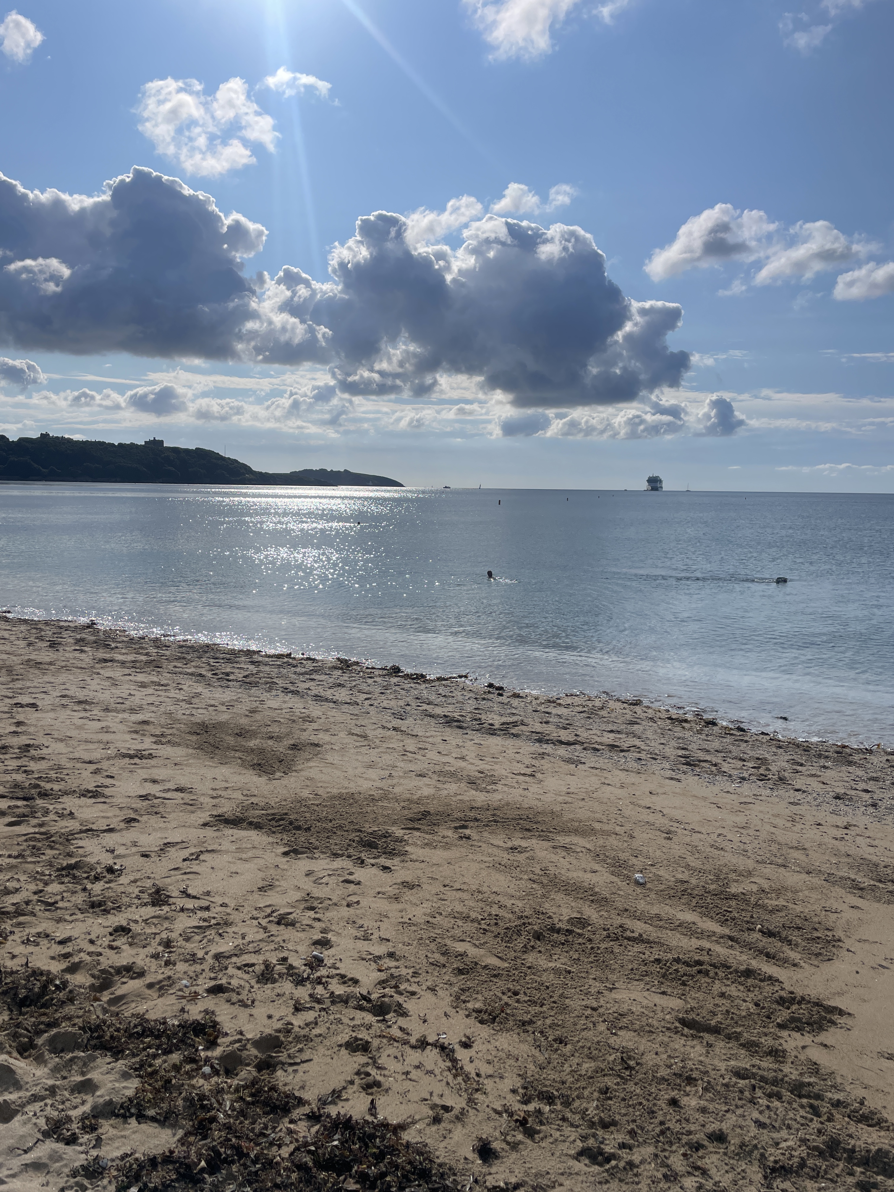 View of Gyllyngvase beach with sun dazzling on sea surface looking towards lighthouse in distance